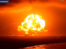 Massive orange fireball explosion on water at night with embers and smoke rising above the horizon