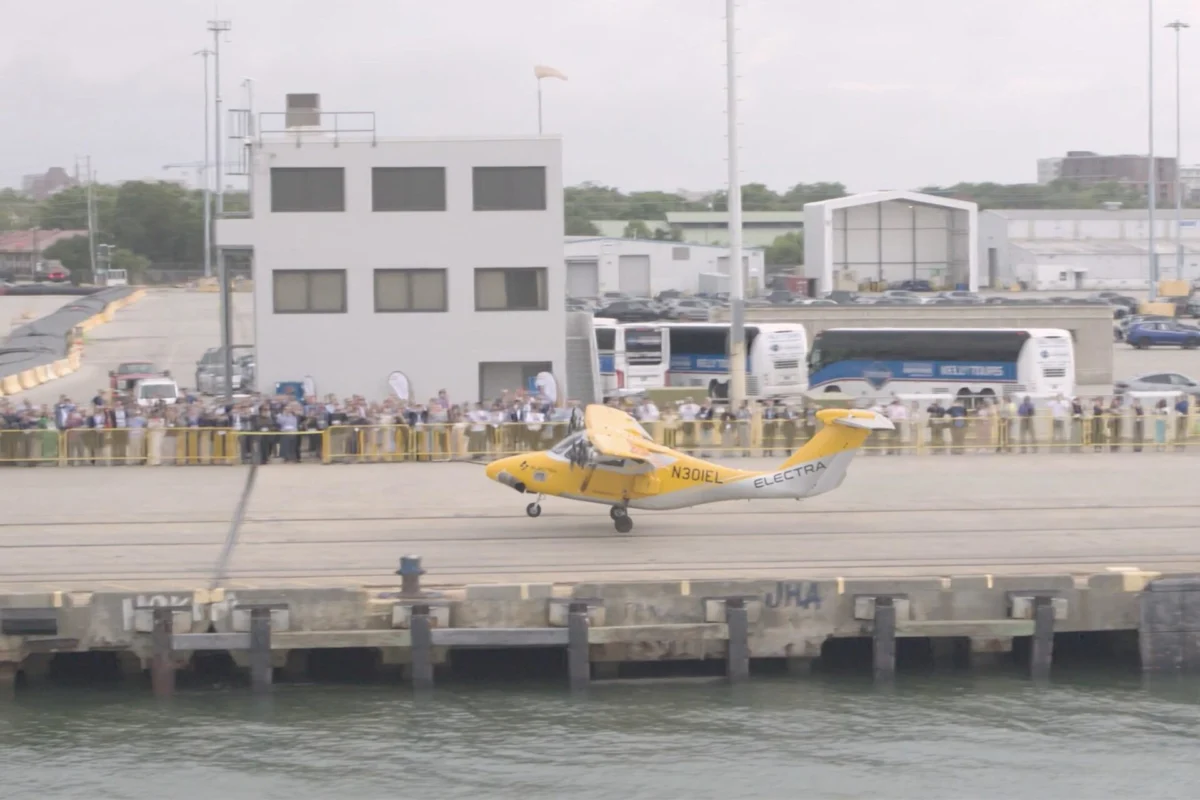 el2 Yellow helicopter lifting off from a waterfront dock while a crowd watches behind barriers