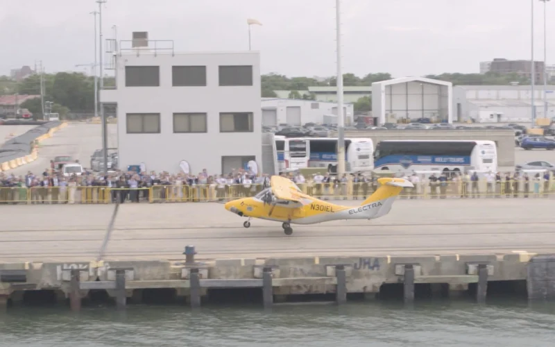 Yellow helicopter lifting off from a waterfront dock while a crowd watches behind barriers