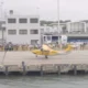 Yellow helicopter lifting off from a waterfront dock while a crowd watches behind barriers