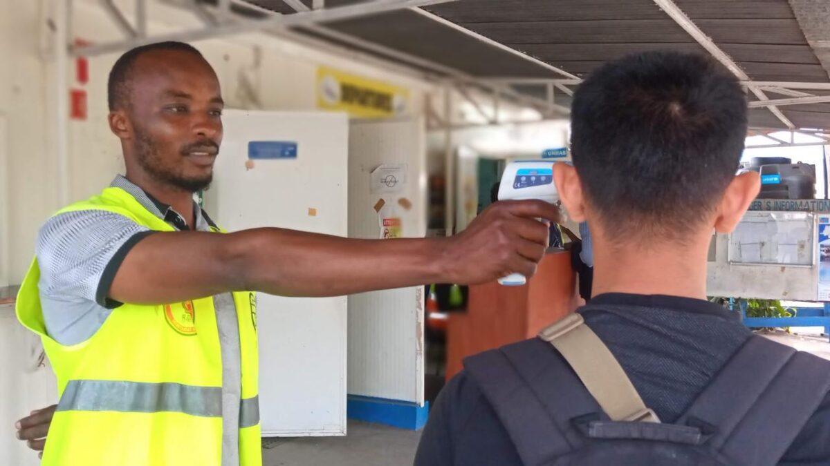 A worker in a bright safety vest uses a handheld thermometer to check a persons temperature at a checkpoint outside a facility