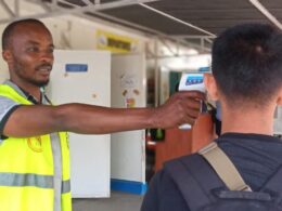 A worker in a bright safety vest uses a handheld thermometer to check a persons temperature at a checkpoint outside a facility