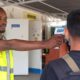 A worker in a bright safety vest uses a handheld thermometer to check a persons temperature at a checkpoint outside a facility