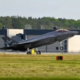 Stealth fighter jet tilting nose up during takeoff on a runway at an airbase with hangars in the background