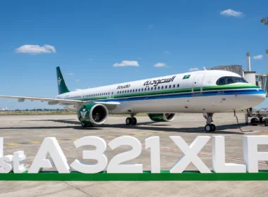Saudia Airbus A321XLR parked at the gate with a green-and-blue stripe livery under a blue sky.Front-right view of a Saudia A321XLR at the jet bridge, white fuselage with green tail and logo.Large '1st A321XLR' signage on the ground in front of the parked airliner on the tarmac.