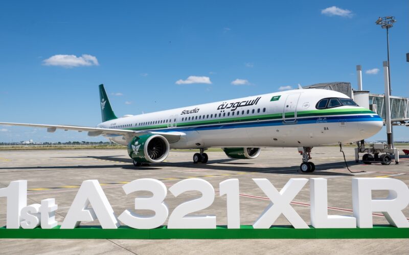 saudia a321xlr Saudia Airbus A321XLR parked at the gate with a green and blue stripe livery under a blue skyFront right view of a Saudia A321XLR at the jet bridge white fuselage with green tail and logoLarge 1st A321XLR signage on the ground in front of the parked airliner on the tarmac