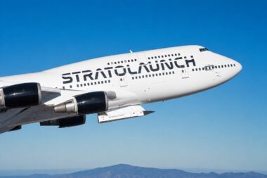 White Stratolaunch aircraft in flight against a clear blue sky with distant mountains on the horizon and its branding visible on the fuselage