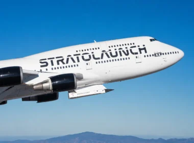 White Stratolaunch aircraft in flight against a clear blue sky, with distant mountains on the horizon and its branding visible on the fuselage