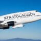White Stratolaunch aircraft in flight against a clear blue sky with distant mountains on the horizon and its branding visible on the fuselage