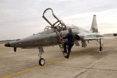 US Air Force fighter jet on a tarmac with the cockpit canopy open and a ground crew member climbing a ladder toward the cockpit