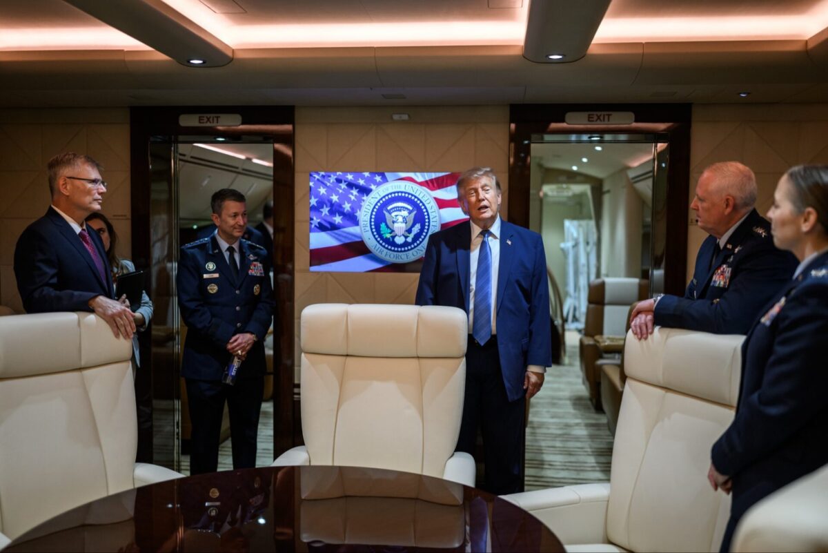 1af1 Man in a blue suit addresses uniformed officers inside a private jet with a screen showing the US presidential seal and flag behind him center stage in the cabin