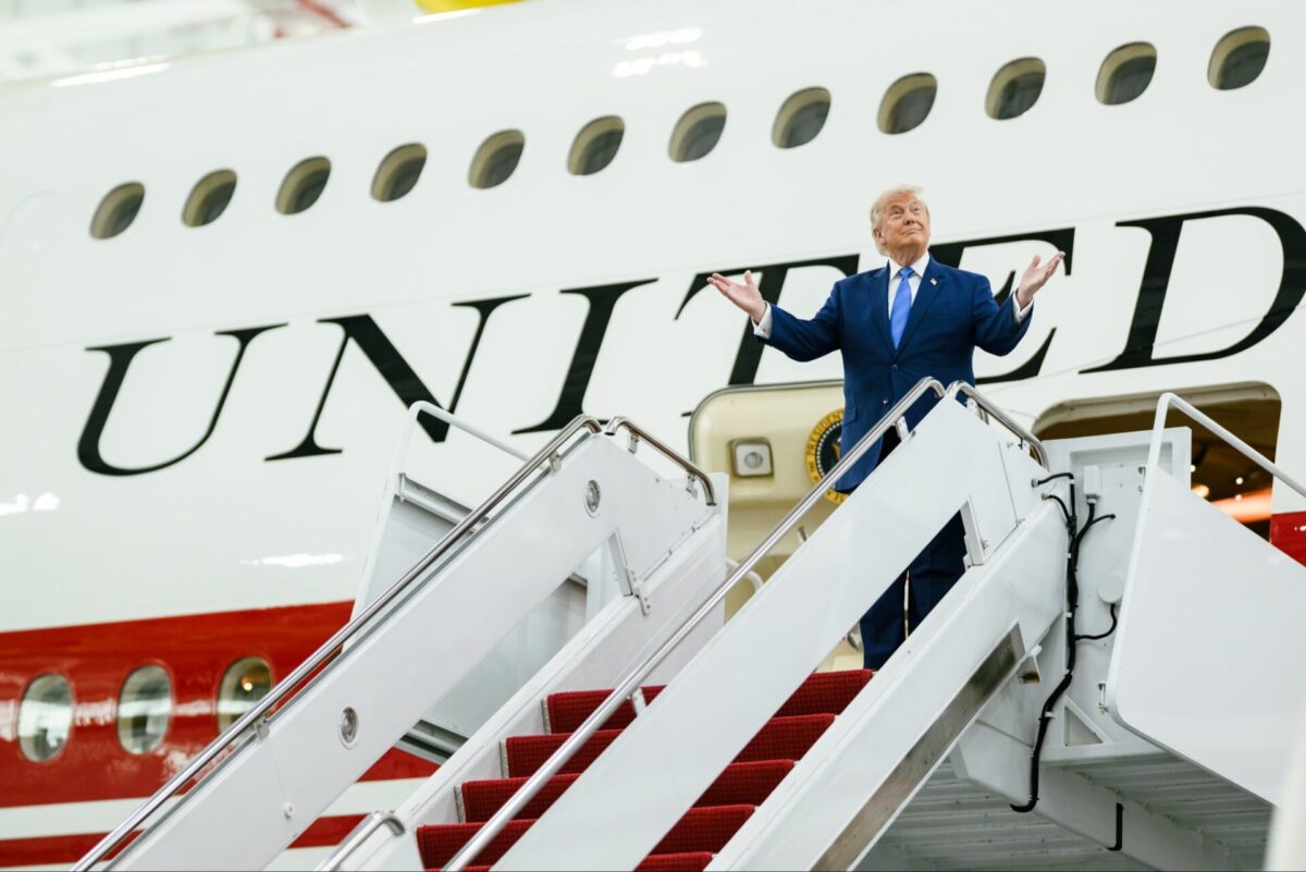 1af2 Former President Donald Trump in a blue suit stands on the aircraft stairs arms open beside a white plane with large UNITED lettering visible behind him