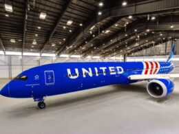 United Airlines jet painted blue parked inside a spacious maintenance hangar with its nose to the left and a visible engine on the right