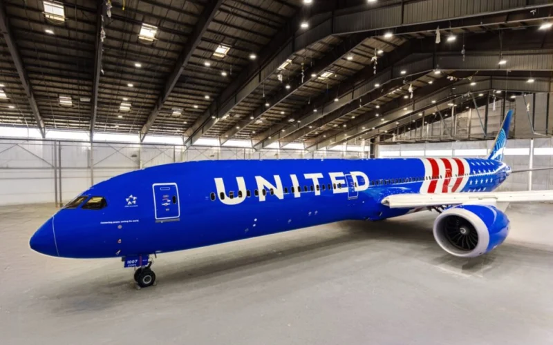 United Airlines jet painted blue parked inside a spacious maintenance hangar with its nose to the left and a visible engine on the right