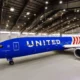United Airlines jet painted blue parked inside a spacious maintenance hangar with its nose to the left and a visible engine on the right