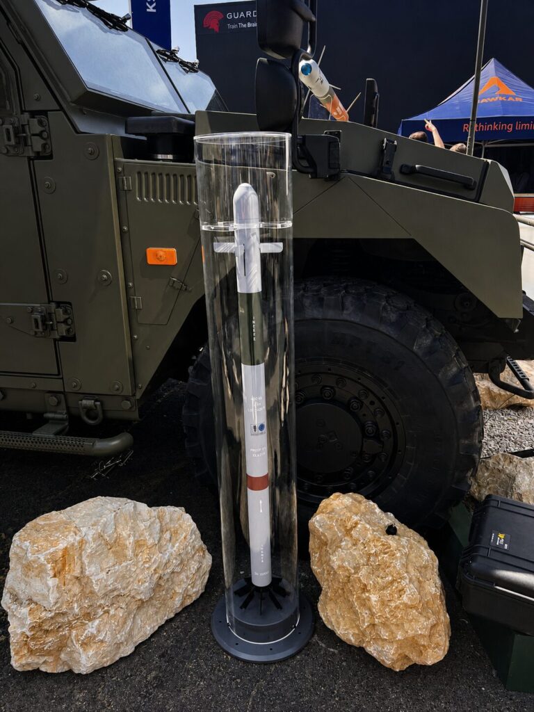 A 70 mm rocket displayed alongside Thales RapidStriker system Missile inside a clear protective case displayed beside an armored military vehicle at an expo site