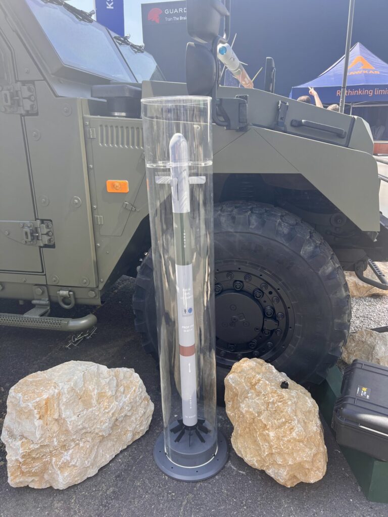 A 70 mm rocket displayed alongside Thales RapidStriker system Clear plastic cylinder enclosing a tall cylindrical device on a stand in front of an armored military vehicle at an outdoor exhibit with rocks around