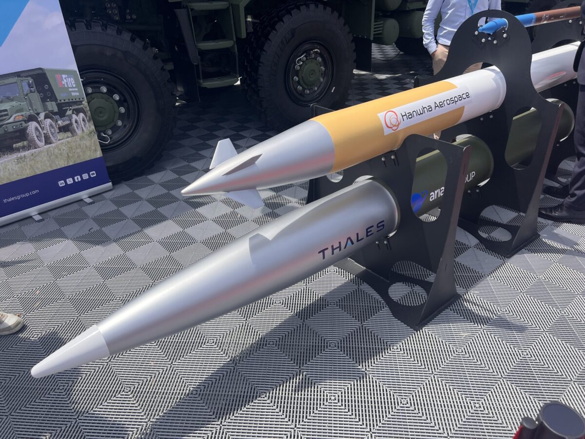 A Hanwha Aerospace CGR 080 rocket mockup displayed alongside Thales FTP 150 munition Missile like weapon mockup on display stand with Hanwha Aerospace branding and a large military truck in the background as an exhibit scene