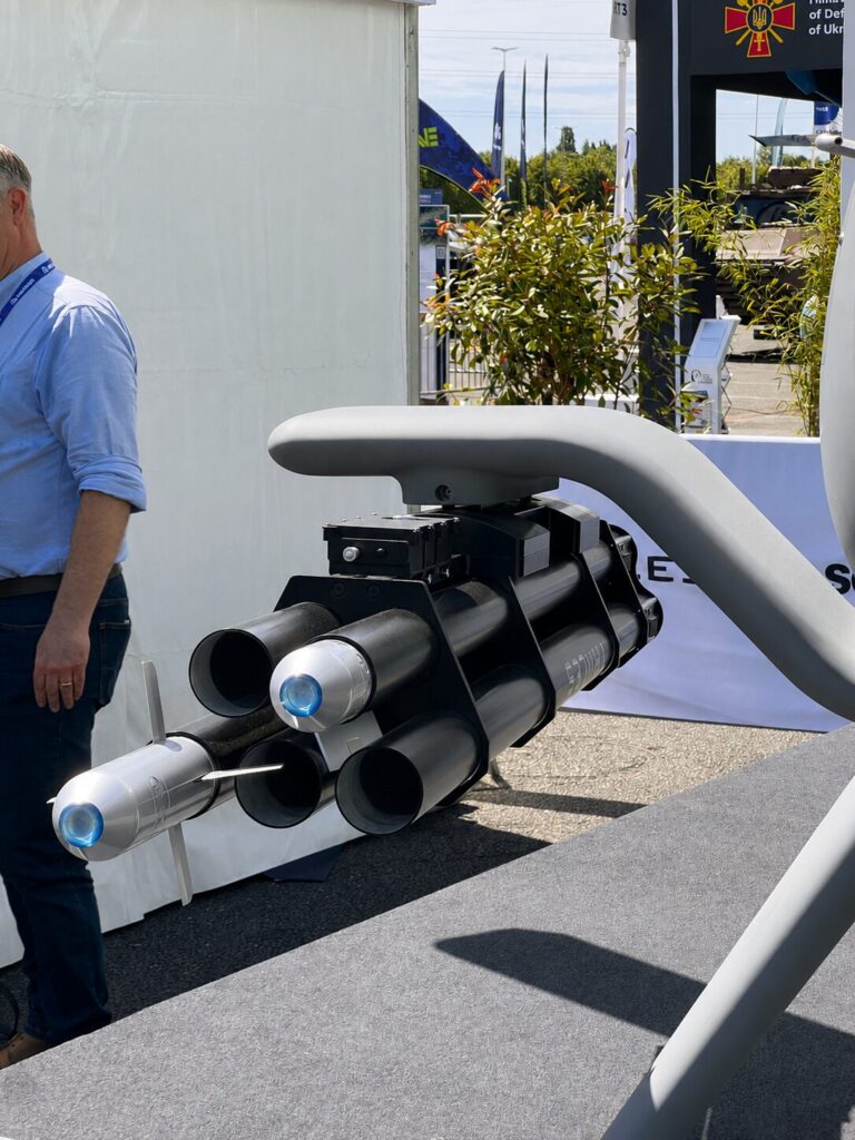 A close up of the Schiebel S 301s side mounted payload station showing Thales LGR 70 mm guided rocket launcher Close up of a multi tube missile launcher mounted on a gray stand at an outdoor defense expo with a man in a blue shirt nearby