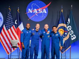 Four astronauts in blue flight suits pose together on stage NASA logo behind them with US flag and agency flags on either side