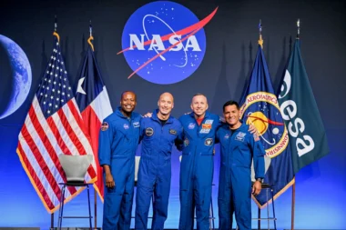 Four astronauts in blue flight suits pose together on stage NASA logo behind them with US flag and agency flags on either side