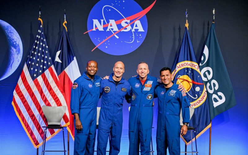 Artemis III crew announcement pillars Four astronauts in blue flight suits pose together on stage NASA logo behind them with US flag and agency flags on either side