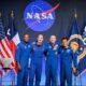 Four astronauts in blue flight suits pose together on stage NASA logo behind them with US flag and agency flags on either side