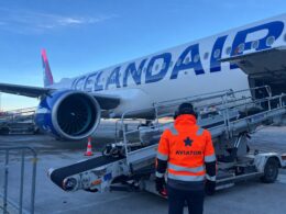 Ground crew member in a bright orange vest stands beside an Icelandair jet on the tarmac with loading stairs at the door and a large engine visible in the wing area