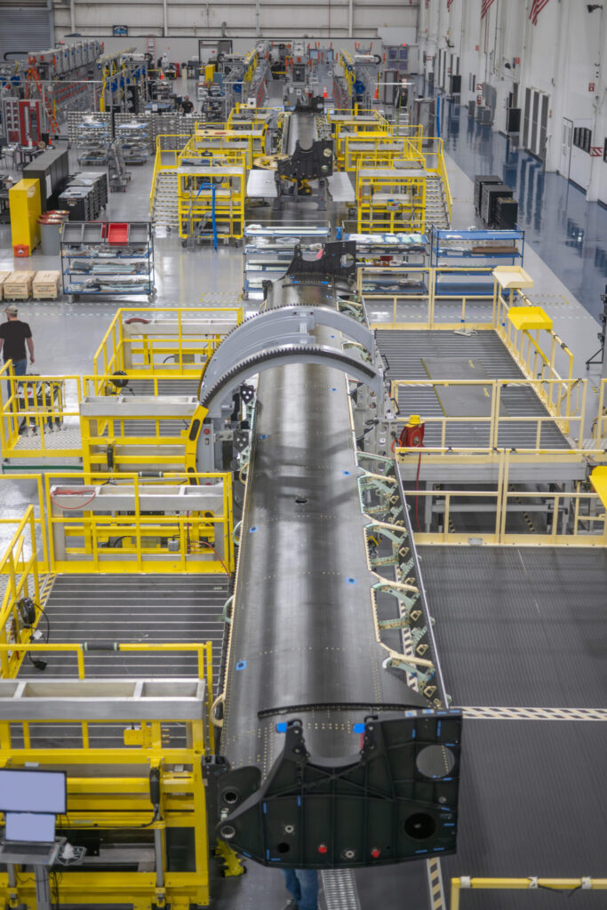 Bell Completes First Two Wing Structures for the MV 75 Cheyenne 1 Aircraft fuselage section on a busy production line with yellow safety rails and industrial machinery in a large factory