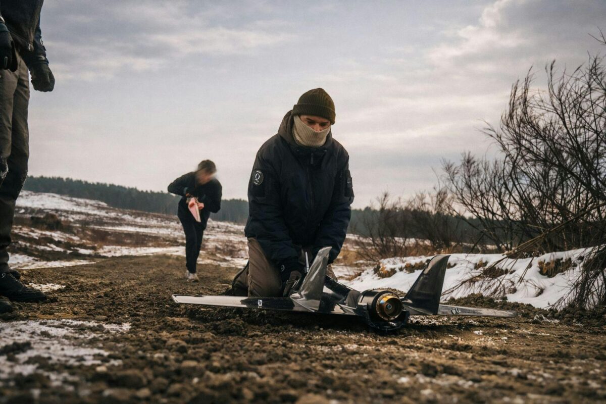 Black bird in Germany during NATO Steadfast Dart exercise in 2026 Person kneels beside a small remote controlled jet on a snowy dirt path others preparing gear in chilly weather