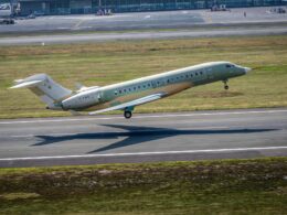 Green private jet taking off on a runway at an airport with grassy field and terminal buildings in the background tail number F WINS