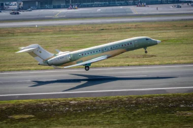Green private jet taking off on a runway at an airport with grassy field and terminal buildings in the background tail number F WINS