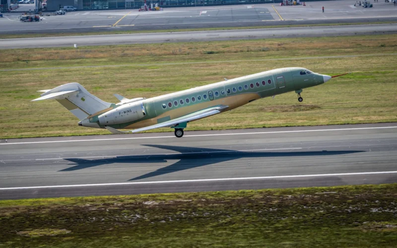 Green private jet taking off on a runway at an airport with grassy field and terminal buildings in the background tail number F WINS