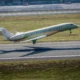 Green private jet taking off on a runway at an airport with grassy field and terminal buildings in the background tail number F WINS
