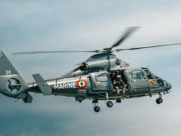 Military helicopter in flight with crew members visible in the open side door gray camouflage and MARINE markings along the fuselage