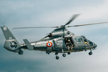 Military helicopter in flight with crew members visible in the open side door gray camouflage and MARINE markings along the fuselage