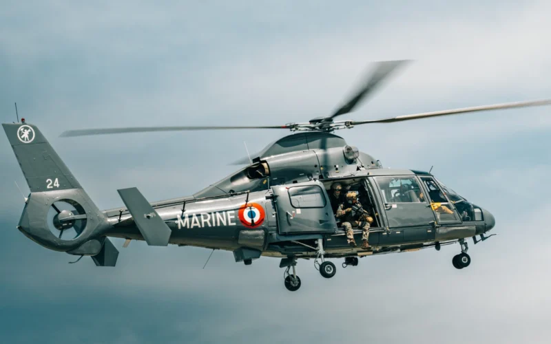 Military helicopter in flight with crew members visible in the open side door gray camouflage and MARINE markings along the fuselage