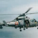 Military helicopter in flight with crew members visible in the open side door gray camouflage and MARINE markings along the fuselage