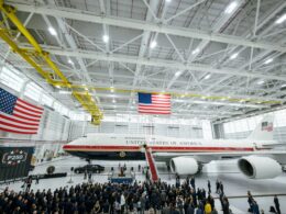 Inside a spacious aircraft hangar a large white jet labeled United States of America sits on the floor as a crowd of people gathers for a ceremony beneath hanging American flags