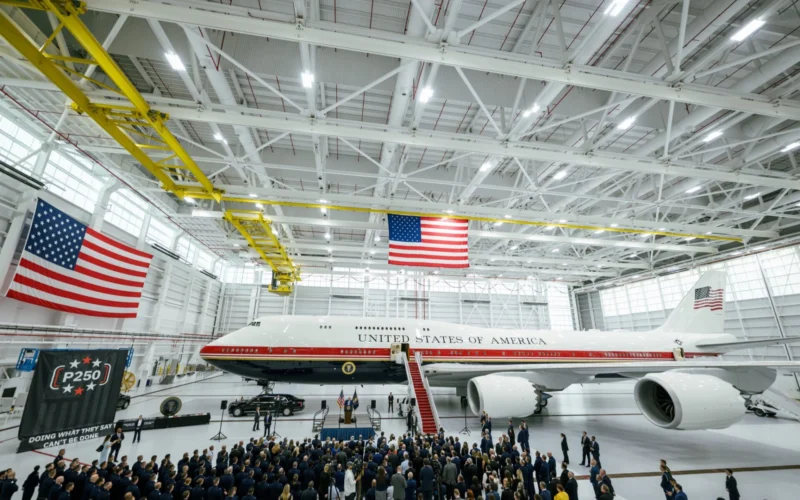 Inside a spacious aircraft hangar a large white jet labeled United States of America sits on the floor as a crowd of people gathers for a ceremony beneath hanging American flags
