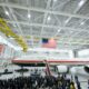 Inside a spacious aircraft hangar a large white jet labeled United States of America sits on the floor as a crowd of people gathers for a ceremony beneath hanging American flags