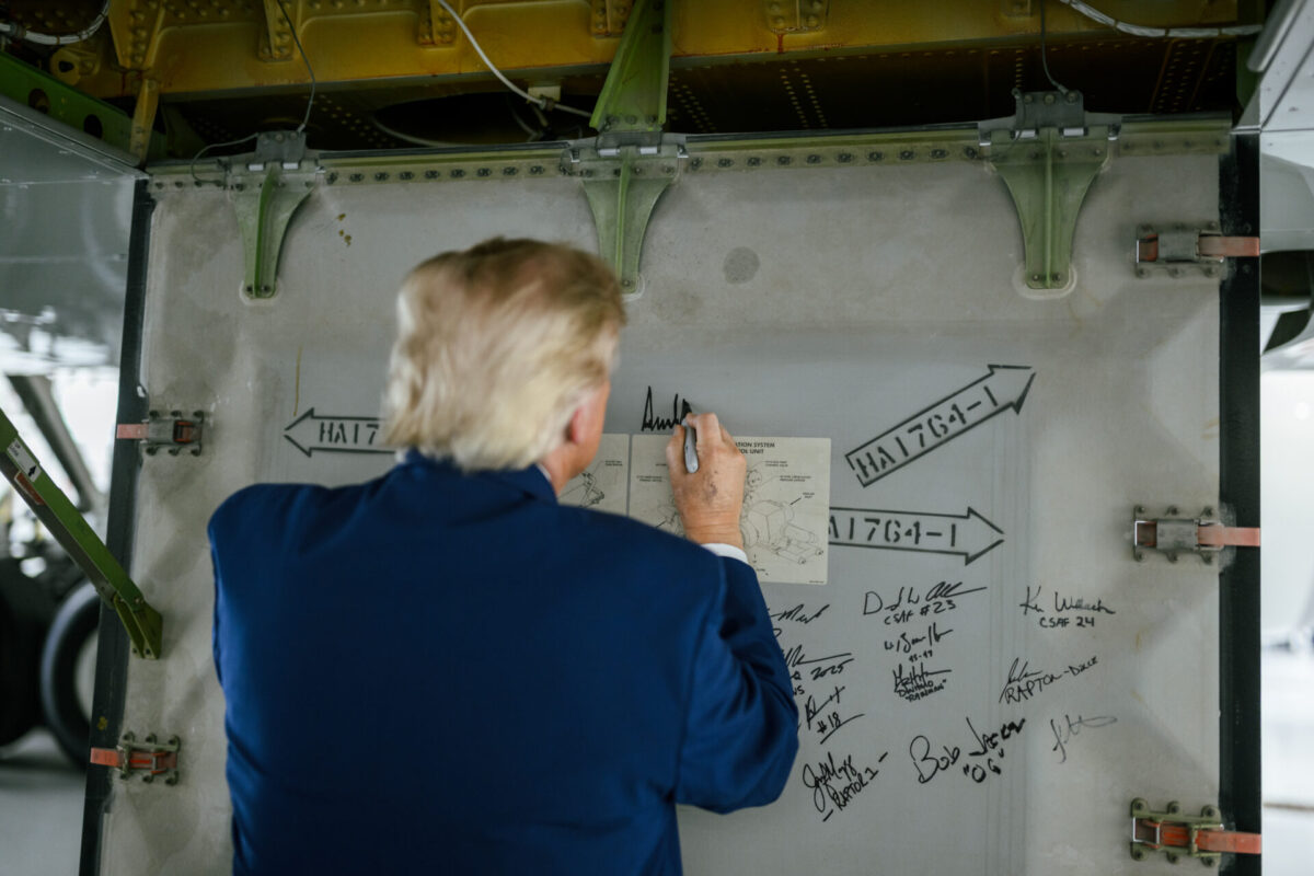 HLQugtVW8AEV8zs Person in a blue jacket signs a metal panel covered with diagrams arrows and many signatures inside a hangar or workshop