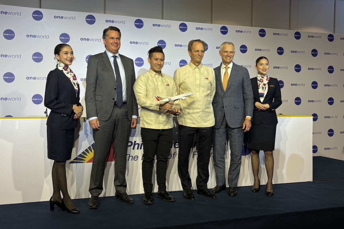 Six people on a stage at an aviation event a man holds a model airplane backdrop shows oneworld logos