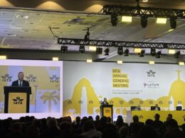 Speaker at a podium on stage during IATA Annual General Meeting with a yellow backdrop and IATA logos visible audience in foreground