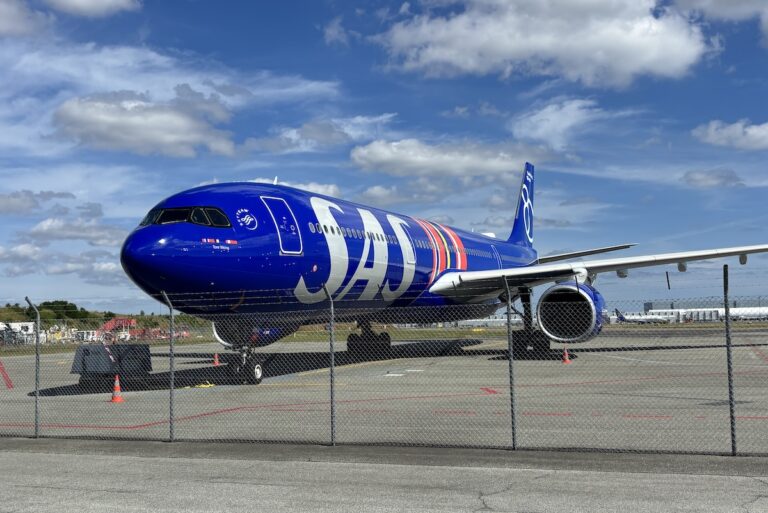Blue commercial jet parked on a taxiway behind a chain link fence under a bright partly cloudy sky engine visible under wing