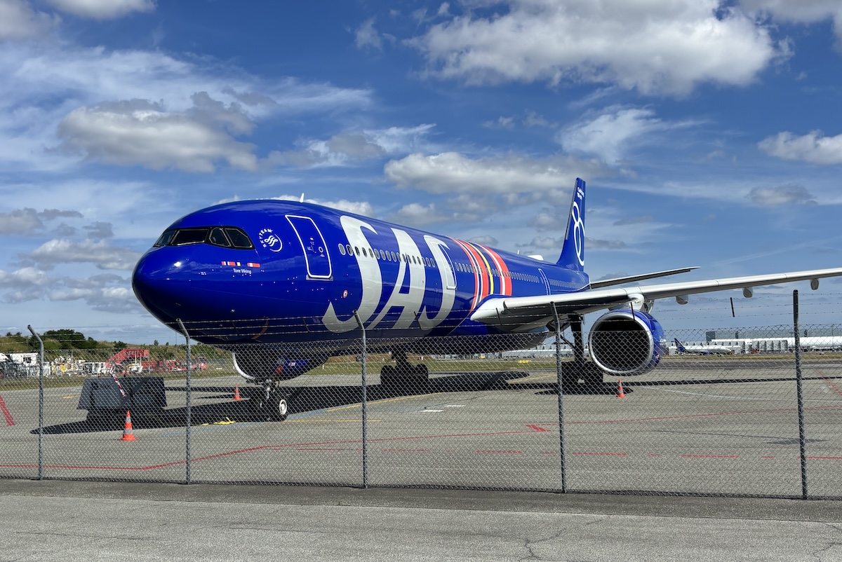 Blue commercial jet parked on a taxiway behind a chain link fence under a bright partly cloudy sky engine visible under wing