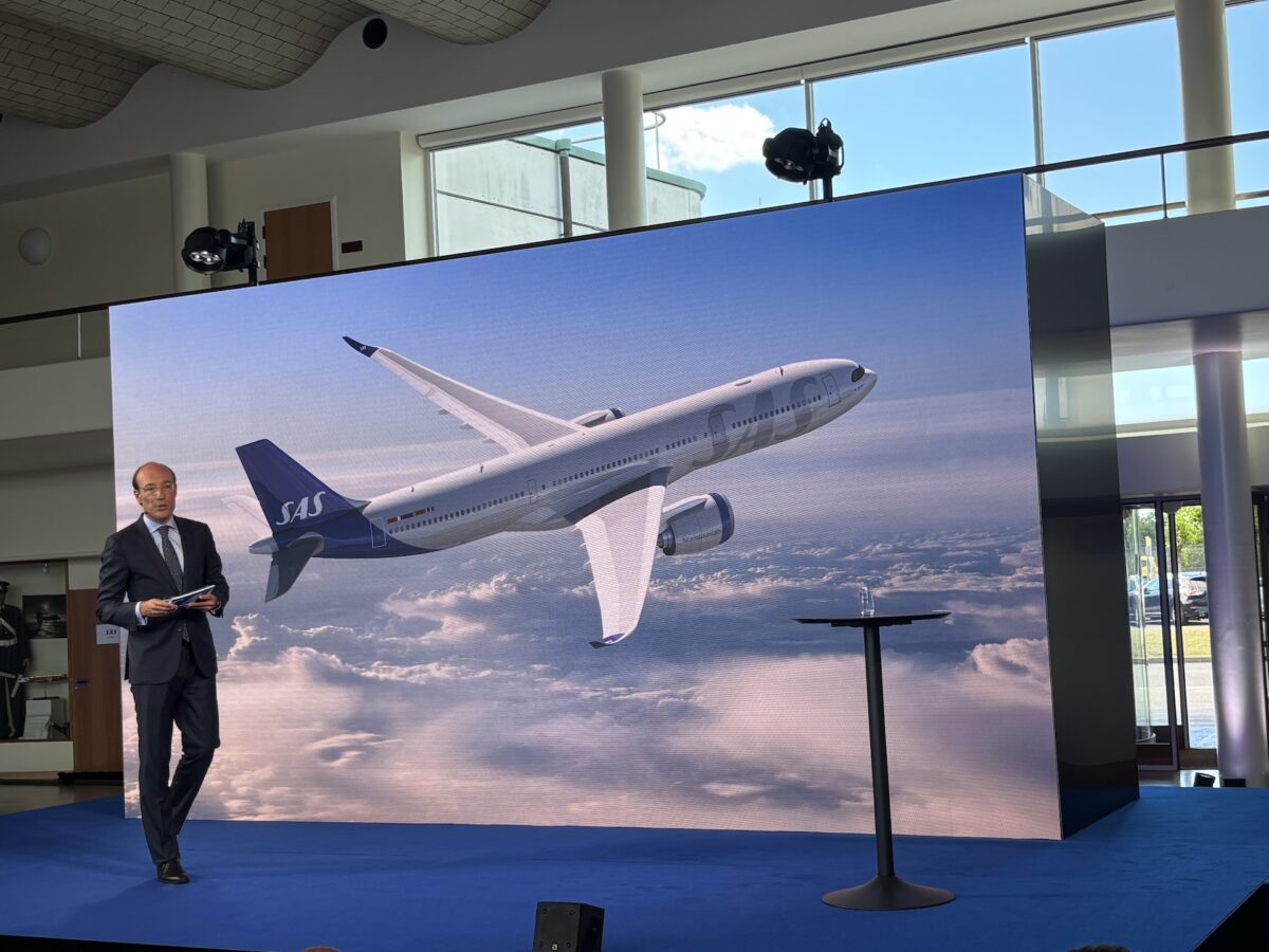 IMG 7179 A presenter in a dark suit stands on a blue carpet in front of a large screen showing a SAS airplane midflight