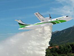 White and green firefighting airplane releasing a thick plume of water over a forested valley with mountains in the background