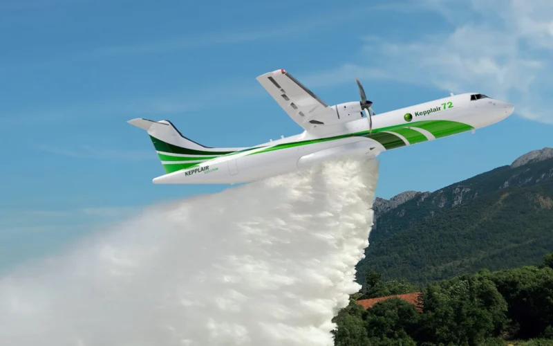 White and green firefighting airplane releasing a thick plume of water over a forested valley with mountains in the background