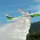 White and green firefighting airplane releasing a thick plume of water over a forested valley with mountains in the background